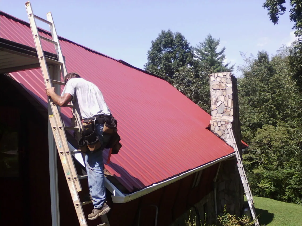 Roofer on a ladder beside a red metal roof and stone chimney — Soffit & Fascia Repair by Roofing Brothers in Waldo